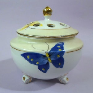 May include: A white ceramic incense burner with a lid, decorated with a blue butterfly design and yellow details. The lid has three holes and a gold finial. The burner stands on three small, gold-trimmed feet.