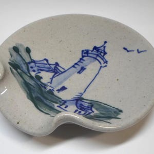 May include: A speckled off-white ceramic spoon rest with a hand-painted blue lighthouse illustration. The design features a tall lighthouse structure, dark green brushstrokes representing land or water, and two small birds in the sky.