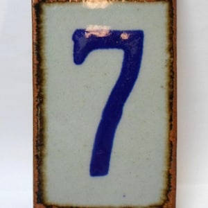 May include: A rectangular ceramic tile featuring the number 7 painted in a deep blue glaze. The tile has a light gray center, a dark brown border, and decorative blue and brown patterns along the top and bottom edges with a glossy finish.