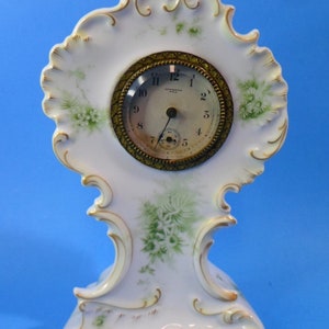 May include: A white porcelain clock with gold accents and a floral design. The clock face is round and has black Roman numeral markings. The clock is on a white background.