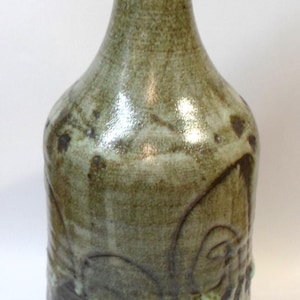 May include: A handcrafted ceramic vase with a narrow neck and rounded body. The vase features a mottled green and brown glaze with textured, organic patterns. The design includes dark brown accents and a light green hue.