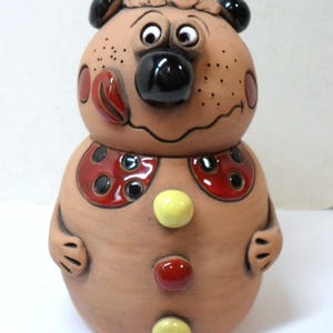 May include: A ceramic figurine of a bear with a brown body, black eyes, and a black nose. The bear is wearing a red and black patterned shirt with yellow buttons.