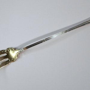 May include: A silver and gold serving fork with a long handle and three prongs. The fork has a decorative design on the handle.