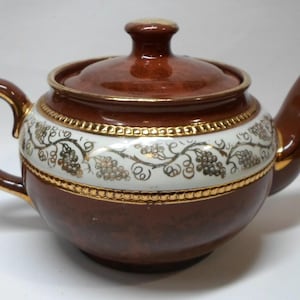 May include: A brown ceramic teapot with a glossy finish, featuring a white horizontal band around the center decorated with a gold-toned grape vine pattern. The teapot includes a matching lid, a curved handle, a spout, and gold-colored trim.