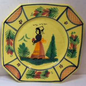 May include: An octagonal yellow ceramic plate featuring a hand-painted illustration of a figure in a dark top and orange skirt standing between green trees. The border is decorated with orange checkered patterns and green floral motifs.