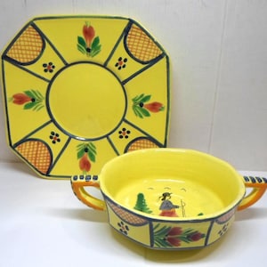 May include: A yellow ceramic bowl and plate set. The bowl has two orange handles and a painted scene inside. The plate is octagonal with a central circle and decorative painted accents in red, green, and black. The set has a folk art style.