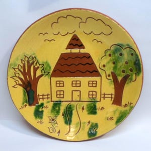 May include: A hand-painted ceramic plate with a yellow background featuring a house, trees, clouds, and a fence. The house has a brown roof and is surrounded by green grass and trees. The plate has a brown rim.