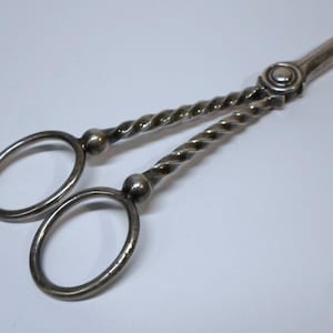 May include: Silver-coloured scissor-like tongs with twisted handles and circular finger holes. The tongs have a decorative design and a hinge at the top. The scissor-like tongs are likely for serving or decorative purposes.
