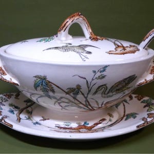 May include: A white ceramic soup tureen with a lid, handles, and a matching saucer. The tureen and saucer are decorated with hand-painted images of birds, plants, and branches in shades of brown, green, and blue. The handle on the lid is brown.