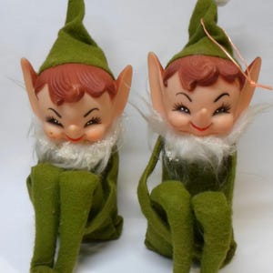 May include: Two identical elf figurines featuring green felt clothing, pointed hats with white pom-poms, and white faux fur collars. Each elf has a moulded plastic face with painted facial features, large pointed ears, and a seated posture.