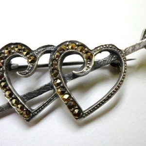 May include: A silver-toned brooch featuring two open hearts pierced by an arrow. The hearts are outlined with small, sparkling stones, and the arrow has a textured design. A classic romantic accessory.