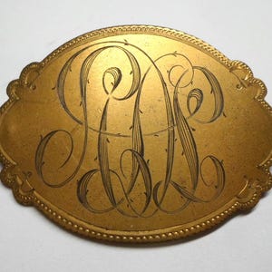 May include: A gold-colored, oval-shaped plaque with a decorative border. The plaque features an ornate, engraved monogram in black script. The design is centered on the plaque and is likely a personalized item.