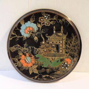 以下が含まれることがあります： 金色の縁が付いた丸い黒い装飾プレート。このプレートは、金色、オレンジ色、青色、緑色の色合いで、塔、花、葉が描かれた、アジア風の詳細なデザインが特徴です。