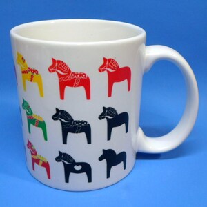 May include: White ceramic mug featuring colorful Dala horse designs. The horses are in various colors, including red, yellow, green, and black. The mug has a curved handle and a smooth, glossy finish. A charming and decorative item for coffee or tea.