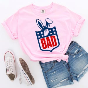 May include: A light pink t-shirt with a graphic design featuring a football, bunny ears, stars, and the word "BAD" in red. The shirt is paired with denim shorts and white sneakers.