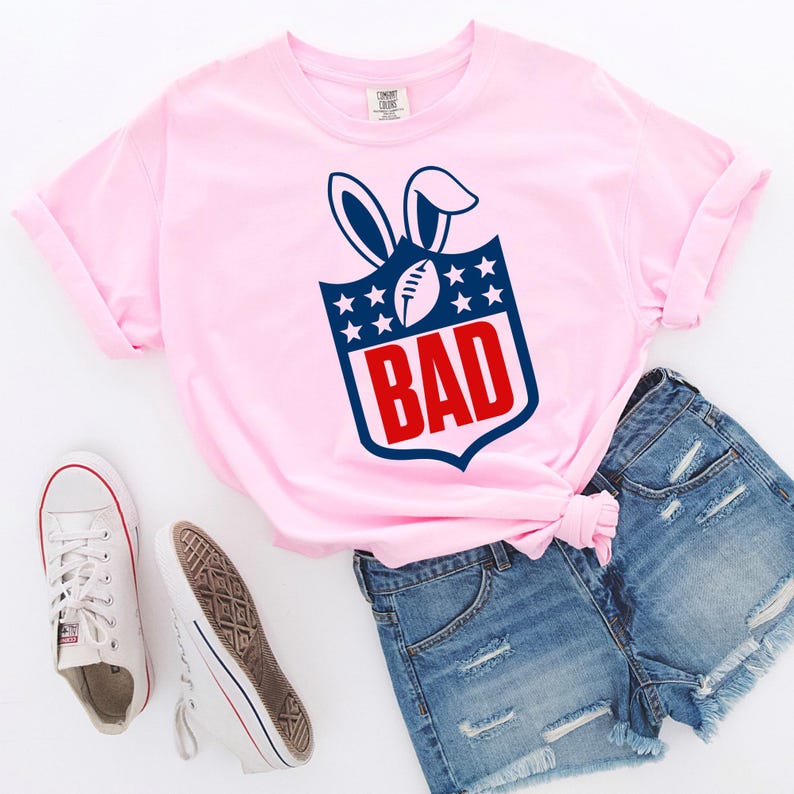 May include: A light pink t-shirt with a graphic design featuring a football, bunny ears, stars, and the word "BAD" in red. The shirt is paired with denim shorts and white sneakers.