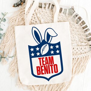 May include: A white canvas tote bag with a blue and red graphic. The graphic features a football, bunny ears, and the words "TEAM BENITO". The bag has two handles.