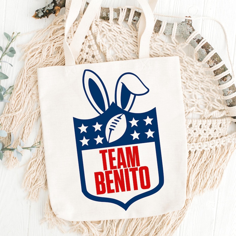 May include: A white canvas tote bag with a blue and red graphic. The graphic features a football, bunny ears, and the words "TEAM BENITO". The bag has two handles.