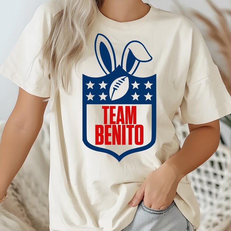 May include: Cream-colored t-shirt featuring a blue and red graphic. The graphic includes bunny ears, a football, stars, and the text "TEAM BENITO" in red. The shirt is a casual, unisex style.