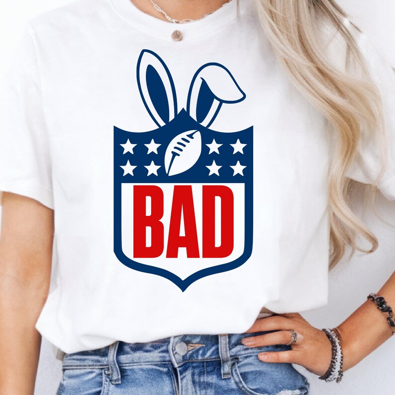 May include: White t-shirt featuring a navy blue and red graphic. The graphic combines a football and bunny ears, with the word "BAD" in bold red letters. The design is set against a navy blue shield with white stars.