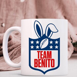 May include: White ceramic mug featuring a blue and white design with a football-shaped object and bunny ears. The text "TEAM BENITO" is in red. The mug has a white handle and is likely for beverages.