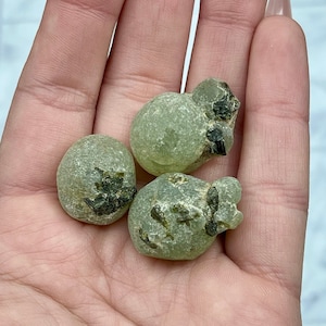 Peut inclure: Trois pierres arrondies vert clair avec des inclusions vert foncé et noires sont tenues dans une main. Les pierres ont une texture légèrement rugueuse et des formes irrégulières. L'arrière-plan est de couleur claire et neutre.