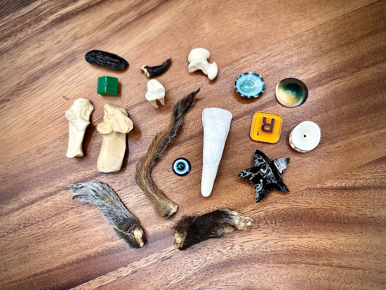 May include: A collection of various animal parts, including bones, fur, claws, and teeth, arranged on a wooden surface. There are also a few decorative items, such as a small green cube, a blue and white eye charm, and a black star-shaped object.