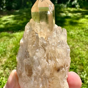 May include: A large, clear quartz crystal cluster with a pointed, clear quartz crystal on top. The cluster has a rough, natural texture and is a light brown color.