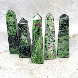 Puede incluir: Cinco puntas de piedra preciosa zultanite verde y rosa, también conocidas como zultanite, están dispuestas en una fila sobre una superficie gris. Las puntas son todas del mismo tamaño y forma, y tienen una variedad de tonos verdes y rosas.