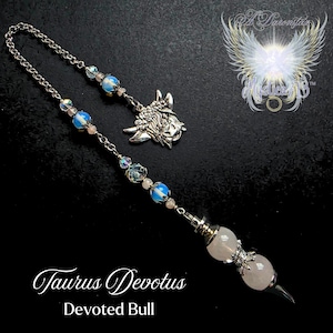 Puede incluir: Un péndulo plateado con una cadena, adornado con cuentas azules y rosas, y un dije de toro. El péndulo tiene un cristal de cuarzo rosa en la parte inferior. El texto "Taurus Devotus" y "Devoted Bull" están en la parte inferior.