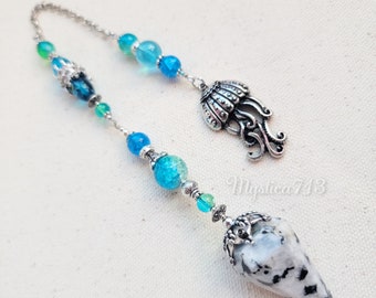 Puede incluir: Un péndulo de obsidiana de copo de nieve blanco y negro con una cadena y cuentas plateadas. La cadena presenta un amuleto de medusa plateado y cuentas de vidrio azules y verdes.