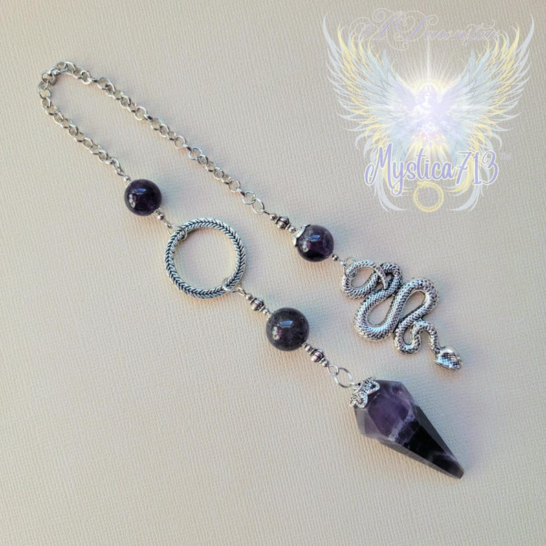 Puede incluir: Un p&eacute;ndulo plateado con cadena, cuentas de amatista y un dije de serpiente. El p&eacute;ndulo presenta una punta de cristal de amatista facetada y un anillo decorativo. El dise&ntilde;o incluye el texto "Mystica713" y un gr&aacute;fico de &aacute;ngel.