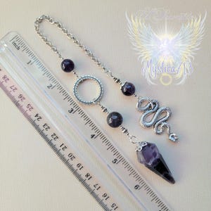Puede incluir: Un p&eacute;ndulo plateado con una cadena, cuentas de amatista y un dije de serpiente. El p&eacute;ndulo tiene una punta de amatista facetada. Una regla est&aacute; colocada junto al p&eacute;ndulo, midiendo aproximadamente 15 cm de largo.