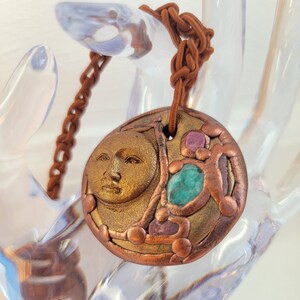 May include: A gold and copper pendant necklace with a moon face design and green and pink gemstones. The necklace is on a brown cord.