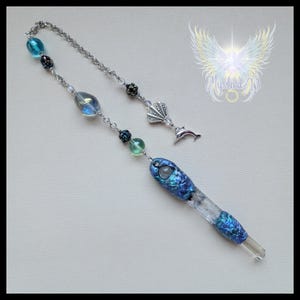 Puede incluir: Una varita de cristal artesanal con un mango azul iridiscente y una punta de cristal transparente. La varita presenta una cadena plateada, cuentas decorativas y un dije de concha y delfín. El diseño incorpora tonos azules y transparentes.