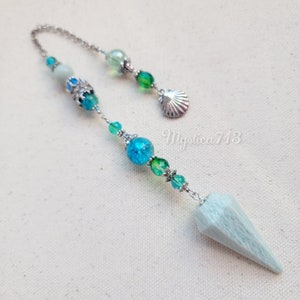 May include: A light blue crystal pendulum with a silver chain and beads. The chain has green, blue, and white beads, and a silver seashell charm. Mystica713 is written on the background.