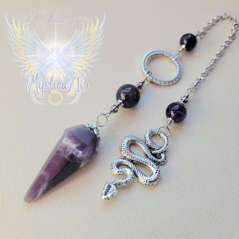 Puede incluir: Un p&eacute;ndulo con una punta de cristal morado y negro, una cadena plateada y un dije de serpiente plateada. La cadena presenta cuentas de piedras preciosas moradas oscuras y un anillo plateado. La imagen tiene el texto "Mystica 713" y un gr&aacute;fico de alas de &aacute;ngel.