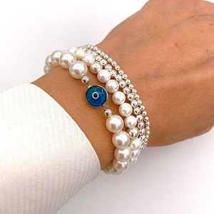 May include: Three bracelets stacked on a wrist. The bracelets include white faux pearls and small silver beads. One bracelet features a blue evil eye charm with a white and black center. The bracelets are worn over a white shirt.