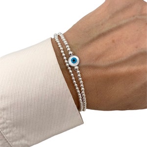 Peut inclure: Deux bracelets de perles argentées sont portés au poignet. L'un des bracelets comporte un charme en forme d'œil bleu et blanc. Les bracelets sont portés sur une chemise blanche.