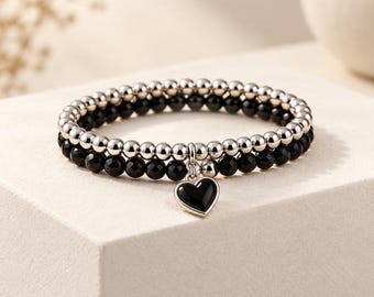 Könnte beinhalten: Ein zweireihiges Perlenarmband bestehend aus einer Reihe polierter silberfarbener Metallperlen und einer Reihe facettierter schwarzer Perlen. Ein herzförmiger Anhänger mit schwarzer Mitte und silberfarbenem Rand hängt in der Mitte des Armbands.