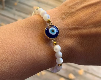 May include: White beaded bracelet with a gold accent and a blue and white evil eye charm. The bracelet is made of small, round, white beads and gold-colored metal accents. The evil eye charm is round with a blue center and a gold border.
