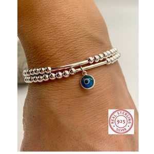 May include: Two silver bracelets with beaded strands and a dangling charm. The charm is a blue and green evil eye design. The bracelets are made of sterling silver.