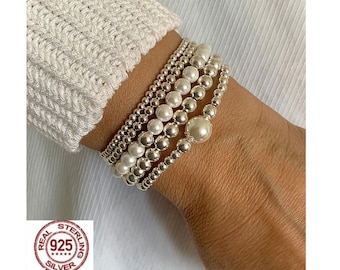 Könnte beinhalten: Fünf Armbänder mit Perlen auf einem Handgelenk. Die Armbänder bestehen aus kleinen Silberperlen und größeren weißen Perlen. Das Bild enthält einen Stempel "925 Silver".
