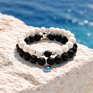 May include: Two stacked beaded bracelets, one black and one white. The bracelets are made of round, textured beads. A silver charm with a blue eye hangs from the bottom bracelet. The background is a blurred view of the sea.