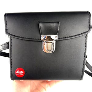 Puede incluir: Funda de cámara de cuero negro con cierre plateado y logotipo "Leitz Wetzlar" rojo y blanco. La funda tiene forma rectangular con cierre de solapa y correa para el hombro. Las costuras son visibles en los bordes.