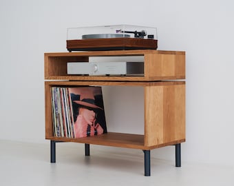 May include: A two-tiered wooden record player stand with black legs. The top shelf has a turntable with a clear lid and a silver amplifier. The bottom shelf has a stack of vinyl records.