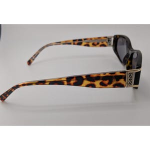May include: Sunglasses featuring a brown and black leopard print pattern on the temples and frame edges. The temples include a metallic silver-toned hinge detail with an embossed S-shaped logo. The lenses are dark and the frame is black.