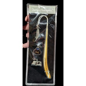 May include: A silver bookmark with a feather, inkwell, and open book charm. The bookmark is curved and has the text "Literature is writ in ink!" on it. The bookmark is packaged in a clear plastic sleeve with a black background and the text "Charming Bookmarks" at the top.