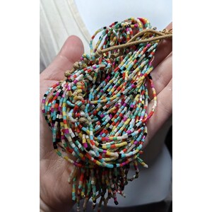May include: A colorful beaded necklace with a multitude of small, rectangular beads in various colors, including red, yellow, black, and blue. The beads are strung together, creating a textured and vibrant accessory. The necklace is held in a hand.