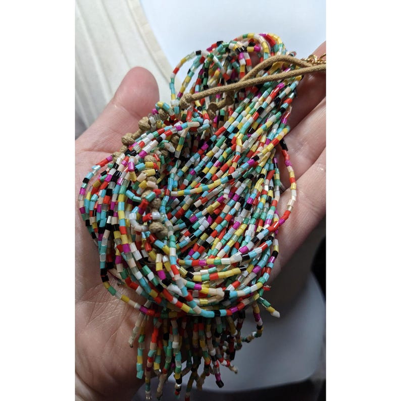 May include: A colorful beaded necklace with a multitude of small, rectangular beads in various colors, including red, yellow, black, and blue. The beads are strung together, creating a textured and vibrant accessory. The necklace is held in a hand.